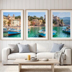 May include: Three framed wall art prints featuring coastal Italian scenes with blue wooden boats floating on turquoise water, colourful Mediterranean buildings, a sandy beach, and a stone castle perched on a cliffside under a clear blue sky.