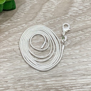 May include: A silver snake chain necklace with a lobster clasp fastening. The chain is coiled and laid flat on a wooden surface.