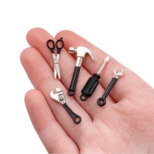Könnte beinhalten: Ein Satz von fünf Miniaturwerkzeugen, darunter ein Hammer, ein Schraubendreher, ein Schraubenschlüssel, eine Schere und ein Schraubenschlüssel. Die Werkzeuge sind silberfarben mit schwarzen Griffen und werden auf einer Hand präsentiert.