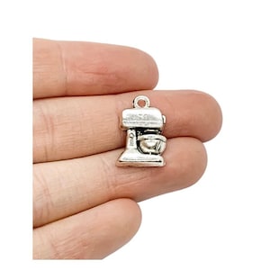 May include: Silver charm in the shape of a stand mixer. The charm is approximately 2.5 cm tall and has a loop at the top for attaching to a necklace or bracelet.