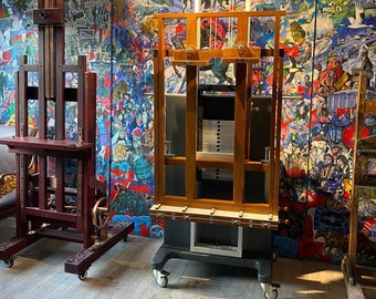 May include: Two wooden artist easels on wheels. One is a dark reddish-brown color, and the other is a lighter brown. The easels are in front of a colorful mural with various figures and scenes. The floor is a light gray wood.