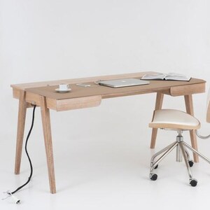 Puede incluir: Un escritorio de madera clara con un cajón incorporado, un portátil y un libro abierto. Una taza de té blanca está sobre el escritorio. Una silla a juego con asiento blanco y patas cromadas está al lado del escritorio.