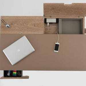 Puede incluir: Vista superior de un escritorio moderno con una superficie marrón claro. Un portátil plateado y un smartphone están sobre el escritorio. El escritorio tiene una sección de madera con un compartimento y una pequeña estantería con material de oficina.