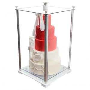 Puede incluir: Una tarta de boda de varios pisos dentro de una vitrina transparente. La tarta tiene capas de glaseado rojo, rosa y blanco, adornada con cintas y perlas. La vitrina es cuadrada con tapa y laterales transparentes y base blanca.