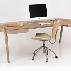Puede incluir: Un escritorio moderno de madera con una superficie marrón claro, un monitor de ordenador, un teclado y un libro. Una silla de madera a juego con asiento color crema y base plateada está colocada frente al escritorio. El escritorio tiene un cajón y un cable de alimentación.