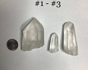 May include: Three clear quartz crystals of varying sizes. The crystals are pointed and have a natural, rough texture. The crystals are arranged on a white surface. #1 - #3