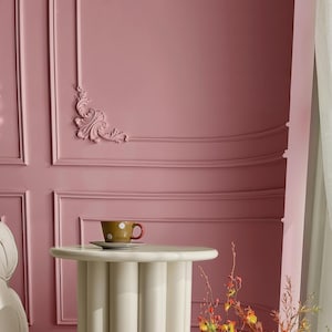 May include: A white side table with a round top and four legs sits in front of a pink wall with decorative molding. A small white saucer with a brown and white polka dot teacup sits on the table.