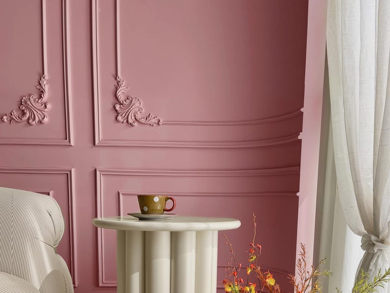 May include: A white side table with a round top and four legs sits in front of a pink wall with decorative molding. A small white saucer with a brown and white polka dot teacup sits on the table.