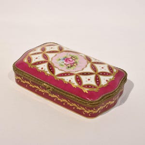 May include: A rectangular porcelain trinket box featuring a deep magenta base with gold filigree accents. The lid displays a white and magenta geometric lattice pattern surrounding a central pink medallion decorated with a painted floral bouquet.