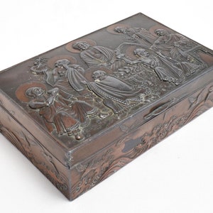 Può includere: Una scatola rettangolare color bronzo con intagli a rilievo dettagliati. Il coperchio presenta una scena con più figure, forse religiose, con aureole. I lati della scatola presentano ulteriori disegni scolpiti, forse floreali o a forma di onda.