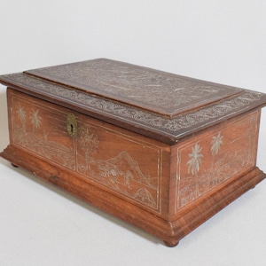 Puede incluir: Una caja de madera con tapa abatible, tallada con intrincados detalles de incrustaciones de plata que representan palmeras y una escena de montaña. La caja tiene una cerradura y una cerradura de latón.