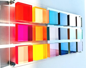 May include: A rectangular clear acrylic wall display featuring three horizontal rows of square, translucent, multi-colored blocks. The blocks are arranged in a grid pattern and secured to the transparent panel with metal hardware.