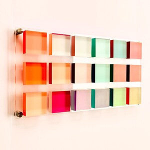 May include: A wall art piece made of 18 acrylic squares in various colors, including orange, pink, green, and purple. The squares are arranged in a grid pattern on a white background.