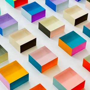 May include: An assortment of colourful, geometric blocks made of different materials. The blocks are arranged in a staggered pattern on a white surface. The blocks are various sizes and shapes, with some being square, rectangular, and triangular.