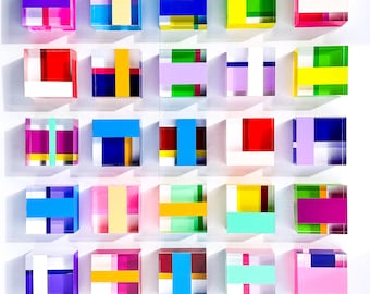 May include: A collection of twenty-five square acrylic blocks arranged in a five-by-five grid. Each block features a unique composition of intersecting translucent and opaque colored stripes in shades of pink, blue, yellow, green, and purple.