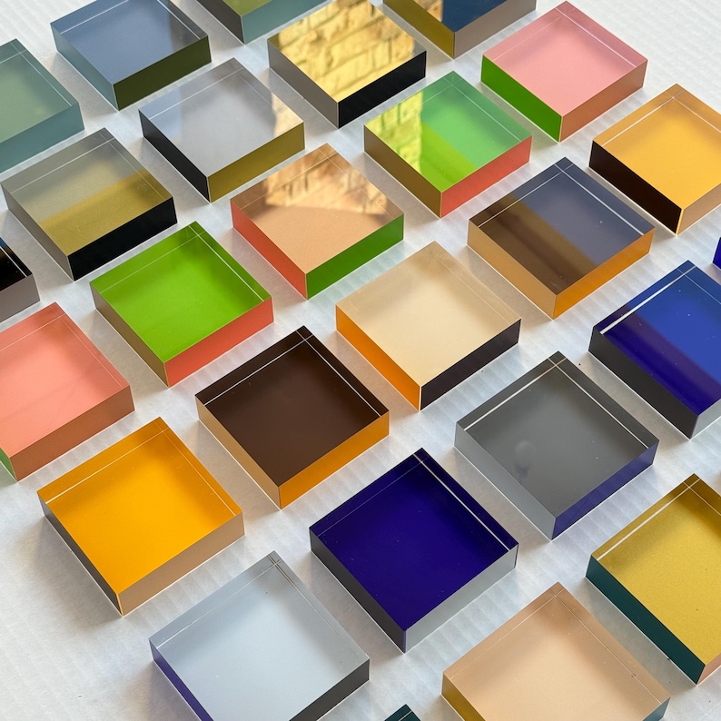 May include: A collection of square acrylic blocks in a range of colours, such as pink, green, yellow, and blue. The blocks are arranged in a grid, highlighting their vibrant hues and reflective surfaces. These blocks are a decorative item.