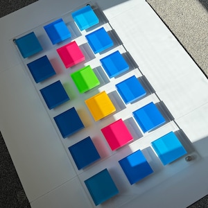 May include: A transparent acrylic panel featuring sixteen coloured square blocks in four rows. The blocks are various shades of blue, pink, green, and yellow. The panel is on a white surface, with sunlight creating shadows.