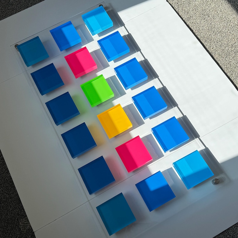 May include: A transparent acrylic panel featuring sixteen coloured square blocks in four rows. The blocks are various shades of blue, pink, green, and yellow. The panel is on a white surface, with sunlight creating shadows.