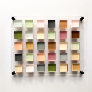 Pode incluir: Um painel de acr&iacute;lico transparente exibe uma grade de 25 cubos transl&uacute;cidos coloridos. Os cubos s&atilde;o dispostos em um padr&atilde;o 5x5, com uma variedade de cores, incluindo rosa, verde, laranja e marrom. Ferragens pretas prendem o painel &agrave; parede.