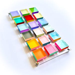 Könnte beinhalten: Ein klarer Acryltablett hält sechzehn farbenfrohe, quadratische Blöcke. Jeder Block zeigt eine andere lebendige Farbe, darunter Schattierungen von Rosa, Gelb, Grün und Blau. Die Blöcke sind in einem Rastermuster angeordnet und erzeugen eine visuell ansprechende Darstellung.