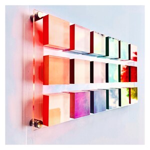 May include: A wall art piece made of 12 acrylic squares in various shades of pink, blue, green, and yellow. The squares are arranged in three rows of four, with each row slightly offset from the one above it. The squares are mounted on a clear acrylic backing.
