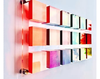 May include: A wall art piece made of 12 acrylic squares in various shades of pink, blue, green, and yellow. The squares are arranged in three rows of four, with each row slightly offset from the one above it. The squares are mounted on a clear acrylic backing.