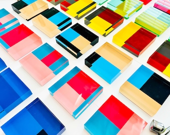 Puede incluir: Una colección de bloques acrílicos geométricos coloridos con varias combinaciones de colores, incluyendo azul, rojo, amarillo, rosa y negro. Los bloques están dispuestos en un patrón de cuadrícula sobre una superficie blanca.