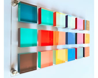 May include: A rectangular clear acrylic wall display featuring three horizontal rows of small, multi-colored square blocks. The blocks are arranged in a grid pattern, showcasing a variety of vibrant hues including red, blue, yellow, and pink.