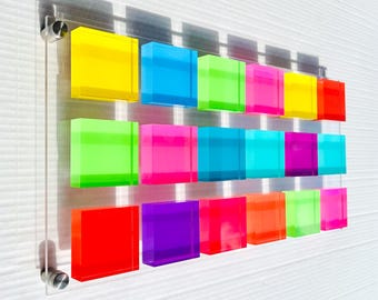 May include: A wall art piece featuring 15 colorful acrylic squares in various shades of yellow, blue, green, pink, red, and purple. The squares are arranged in a grid pattern and are mounted on a clear acrylic panel.