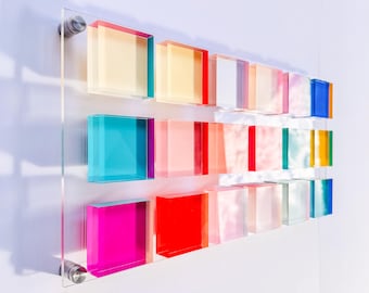 Puede incluir: Una moderna obra de arte mural con un panel acrílico transparente y tres filas de bloques cuadrados coloridos y translúcidos. Los bloques muestran una gama de colores, incluyendo rosa, rojo, azul, amarillo y blanco, creando un diseño vibrante.