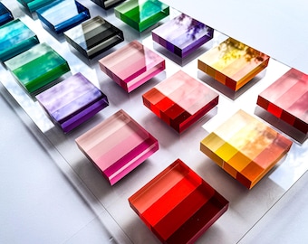 Puede incluir: Un tablero de exhibición de acrílico transparente con una cuadrícula de bloques cuadrados translúcidos multicolores. Los bloques están dispuestos en filas y columnas, mostrando una variedad de tonos vibrantes que incluyen rojo, naranja, amarillo, rosa, morado, azul, verde y gris.