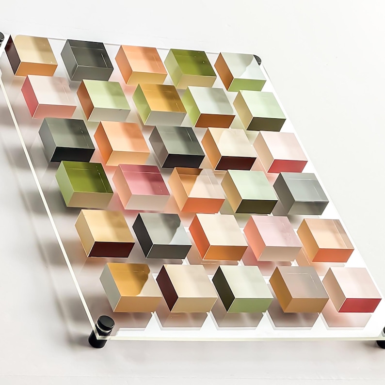 Pode incluir: Uma obra de arte moderna com um painel de acr&iacute;lico transparente com uma s&eacute;rie de cubos transl&uacute;cidos coloridos. Os cubos est&atilde;o dispostos em um padr&atilde;o de grade, mostrando uma variedade de cores, incluindo rosa, verde, laranja e cinza. A obra de arte &eacute; montada com ferragens pretas.