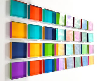 May include: A wall-mounted art installation featuring rows of colorful, translucent square blocks. The blocks display a spectrum of colors, including turquoise, orange, yellow, blue, green, and purple. The artwork creates a vibrant and modern aesthetic.
