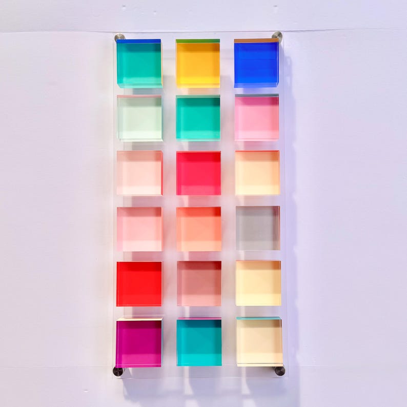 May include: A clear acrylic wall art piece featuring sixteen colourful square blocks in shades of blue, green, yellow, pink, red, and peach. The blocks are arranged in a grid pattern, mounted on a clear backing.