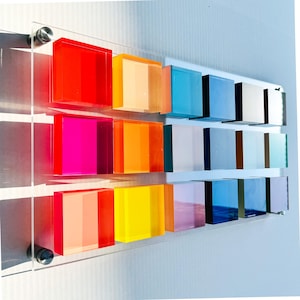 May include: A wall-mounted display featuring a clear acrylic panel secured by four silver metal standoffs. Attached to the panel are fifteen translucent, square-shaped blocks arranged in three rows, showcasing a vibrant spectrum of colors.