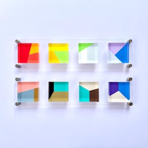May include: A set of eight acrylic squares with geometric patterns in various colors, including red, yellow, green, blue, purple, and brown. The squares are mounted on a white wall.