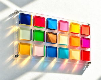 Puede incluir: Un panel rectangular de pared con dieciocho azulejos cuadrados de acrílico translúcido y multicolor dispuestos en cuadrícula. Las piezas muestran un espectro de colores que incluye azul, rojo, verde, amarillo, naranja, rosa y marrón, montadas sobre una placa transparente.