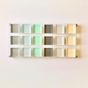 May include: A clear acrylic display with sixteen square glass tiles in various pastel colors, including gray, green, and beige. The tiles are arranged in a grid pattern and mounted with brass hardware.