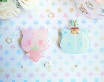 May include: Two pastel-colored cat-shaped pins with cute faces. One pin is pink with a strawberry and the other is blue with a green plant.