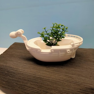 Puede incluir: Una maceta blanca en forma de barco con un diseño decorativo, que contiene una pequeña planta verde. La maceta tiene un frente curvo y una barandilla detallada. La planta añade un toque de naturaleza.