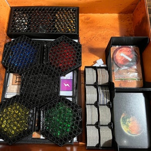 May include: An open box with compartments for storing game pieces. The compartments are filled with small, colorful plastic pieces, cards, and a small bag. The box is made of wood and has a brown finish.