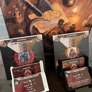 May include: A tabletop fantasy game setup with game cards, card holders, and a game board. The cards feature illustrations of fantasy creatures and game mechanics. The board shows a map and various game pieces.