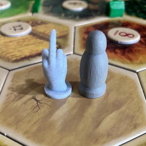 Puede incluir: Dos piezas de juego azul claro, una con forma de mano haciendo una peineta y la otra una figura redondeada, están sobre un tablero de juego hexagonal. El tablero presenta varias fichas de terreno y fichas numéricas.