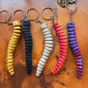 Op de afbeelding: Vijf gearticuleerde, gesegmenteerde, wormachtige sleutelhangers in geel, zwart, gebroken wit, oranje-rood en een paars-blauw verloop. Elke sleutelhanger heeft een metalen ring en ketting, waarvan er één is bevestigd aan een zilverkleurige huissleutel.