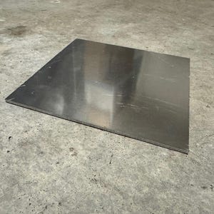May include: A rectangular, silver-colored metal sheet. The sheet has a brushed finish and is lying on a concrete surface. The edges of the sheet are clean and straight. The metal reflects the light.