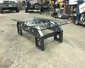 Puede incluir: Una mesa con tapa de vidrio y estructura de metal. La estructura es de metal grueso de aspecto industrial con agujeros y tornillos. La mesa está en un taller.