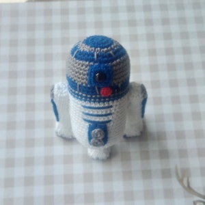 May include: A handmade crochet R2-D2 droid figure. The figure is primarily white with blue and gray accents. It features a black eye, a red tongue, and small gray buttons. The droid is a whimsical representation of the Star Wars character.