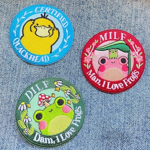 May include: Three embroidered iron-on patches with frog designs. The first patch is blue with a yellow duck and the text "CERTIFIED QUACKHEAD". The second patch is red with a pink frog and the text "MILF Man, I Love Frogs". The third patch is green with a green frog and the text "DILF Dam, I Love Frogs".