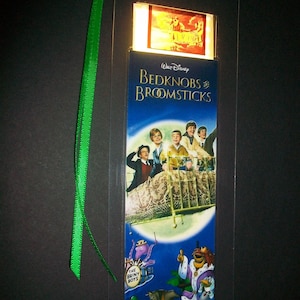 May include: A clear plastic bookmark featuring the movie poster for Walt Disney's "Bedknobs and Broomsticks." The bookmark has a green ribbon attached and includes colorful illustrations of characters and scenes from the film.