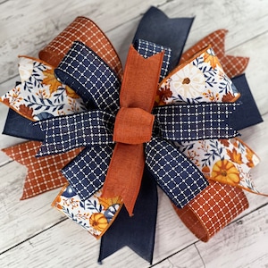 May include: A large, decorative bow made with four different ribbons. The ribbons are a navy blue with a white grid pattern, a rust orange, a white with a floral pattern, and a rust orange with a white grid pattern. The bow is tied with a large rust orange bow in the center.